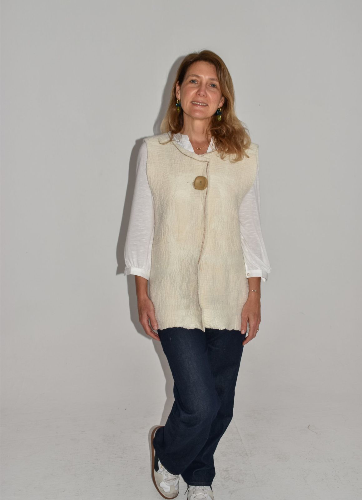 Mujer de pie con un chaleco largo de nuno fieltro marfil, cerrado con un solo botón de madera, sobre camisa blanca, jean oscuro recto y zapatillas.