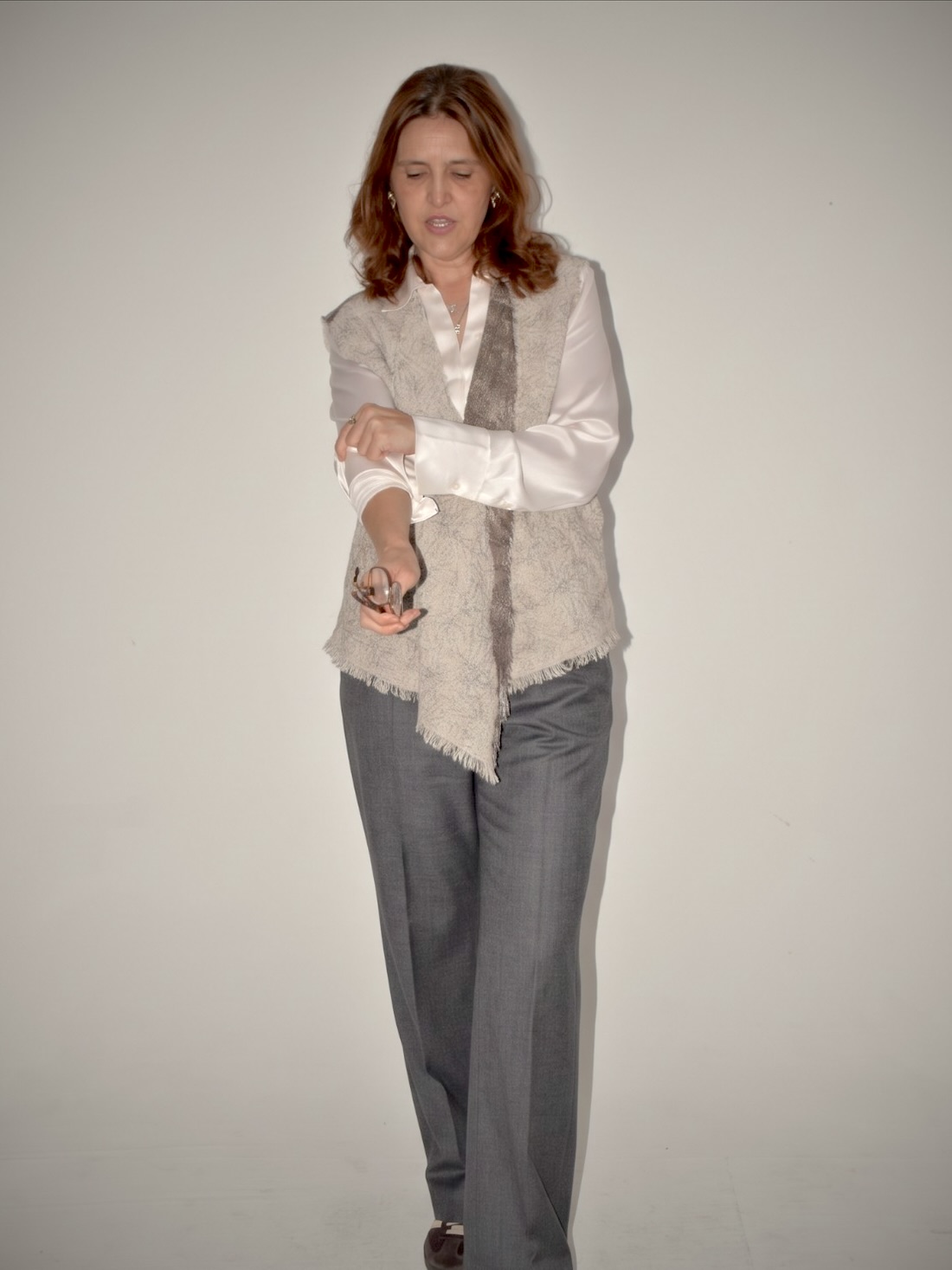 Mujer caminando con un chaleco largo de nuno fieltro gris claro con flecos y una franja marrón, sobre camisa blanca de seda y pantalón sastrero gris.