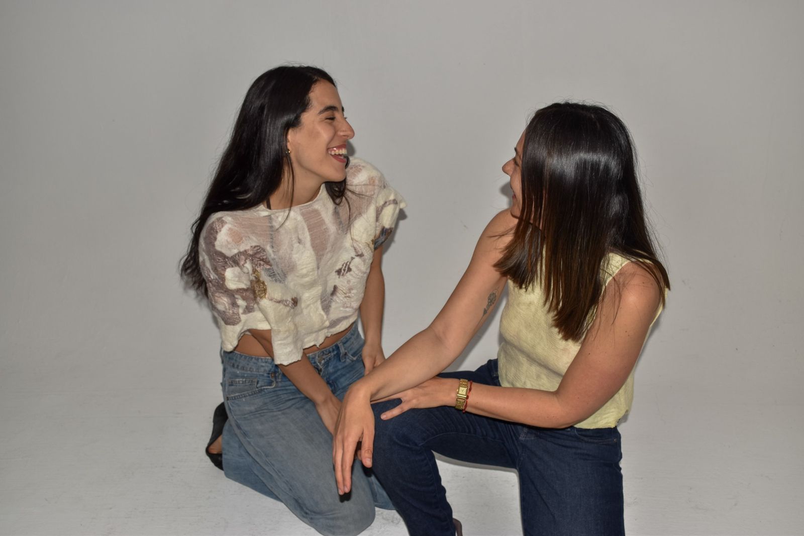 Dos mujeres arrodilladas riéndose frente a frente. Una lleva una prenda corta de gasa y lana marfil con trazos castaños; la otra, una musculosa de lana amarillo pálido.