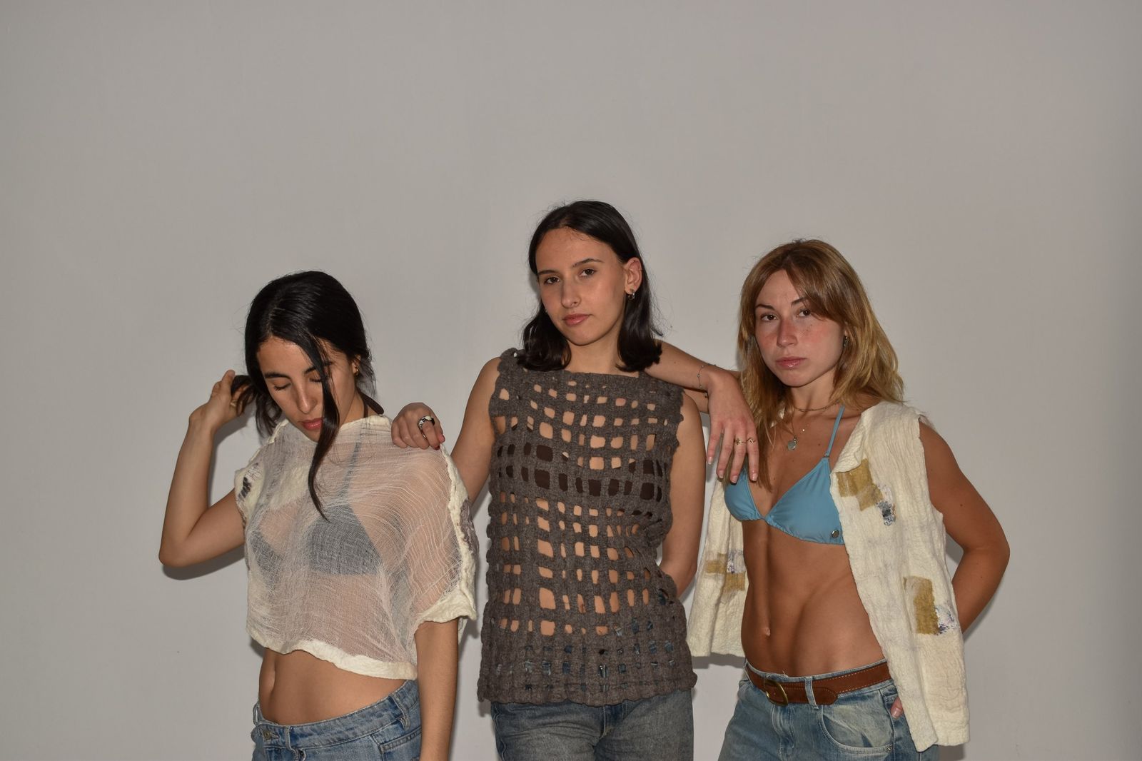 Tres mujeres de pie, apoyadas entre sí, con una prenda de gasa marfil, una musculosa calada de lana gris y una camisa marfil abierta.